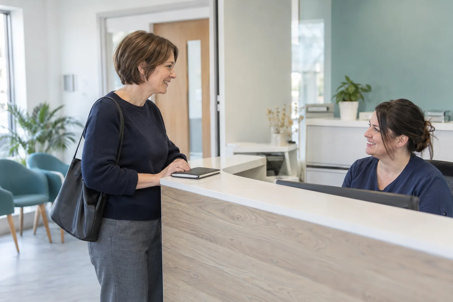 Woman at a dental practice reception asking about NHS dental appointments, with a closed notebook beside her.